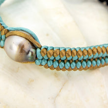 Load image into Gallery viewer, A close-up of a handmade braided cord bracelet featuring light blue and tan woven cords, a large iridescent pearl bead, and a gold accent cord, resting on a light marble surface.