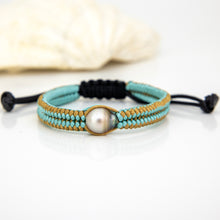 Load image into Gallery viewer, A handmade macrame bracelet featuring light blue and tan woven cords, a central round pearl, and black adjustable sliding knots, displayed on a white surface with a blurred seashell in the background.