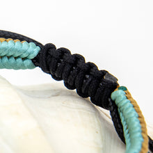 Load image into Gallery viewer, Close-up of a woven cord bracelet or necklace detail with black braided cords and light aqua and tan accents, resting on a white surface against a white background.