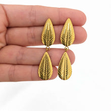 Load image into Gallery viewer, A pair of gold leaf drop earrings held between fingers against a plain white background.