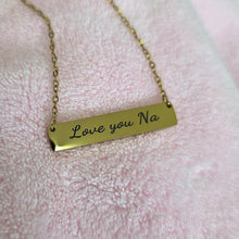 Load image into Gallery viewer, A gold bar necklace with the engraved text 'Love you Na' resting on a soft pink textured fabric background.