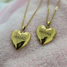 Load image into Gallery viewer, Two gold heart-shaped locket necklaces on chains, one engraved with 'PRASAD' and the other with 'PRIYONA', displayed on a soft pink textured fabric background.