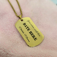 Load image into Gallery viewer, A gold-colored metal dog tag necklace with customizable text, resting on a soft pink textured fabric background.