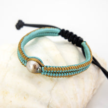Load image into Gallery viewer, A handmade macrame bracelet with light blue and tan woven cords, a silver-toned round bead, and a black adjustable cord, displayed on a white seashell.
