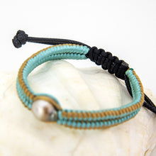 Load image into Gallery viewer, A handmade adjustable bracelet with a light blue and tan braided cord, a black knot section, and a silver bead closure, resting on a white stone.
