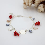 A silver bracelet with red coral and mother of pearl beads, featuring a frangipani charm, displayed alongside a piece of coral.