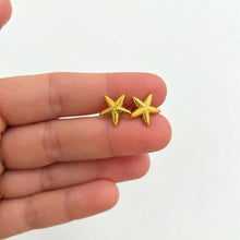 Load image into Gallery viewer, A pair of small gold starfish stud earrings held between a person's fingers against a plain light background.
