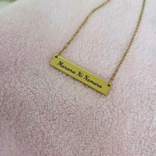 Load image into Gallery viewer, A gold bar necklace with the words 'Merama Ni Namara' engraved on it, resting on a soft pink textured fabric background.
