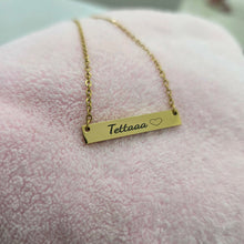 Load image into Gallery viewer, A gold bar necklace with the name 'Tetaaaa' and a small heart engraved on it, resting on a soft pink textured fabric background.
