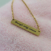 Load image into Gallery viewer, A gold bar necklace engraved with the phrase 'through every season' in black script, resting on a soft pink textured fabric background.
