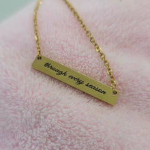 Load image into Gallery viewer, A gold bar necklace engraved with the phrase 'through every season' in black script, resting on a soft pink textured fabric background.
