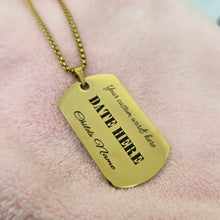 Load image into Gallery viewer, A gold-colored dog tag necklace with customizable text, resting on a soft pink textured fabric background.
