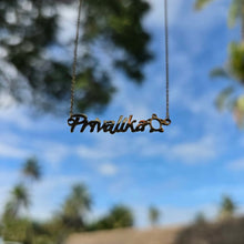 Load image into Gallery viewer, A gold nameplate necklace spelling 'Pravalika' with a turtle accent, suspended against a blurred outdoor background of blue sky and green foliage.
